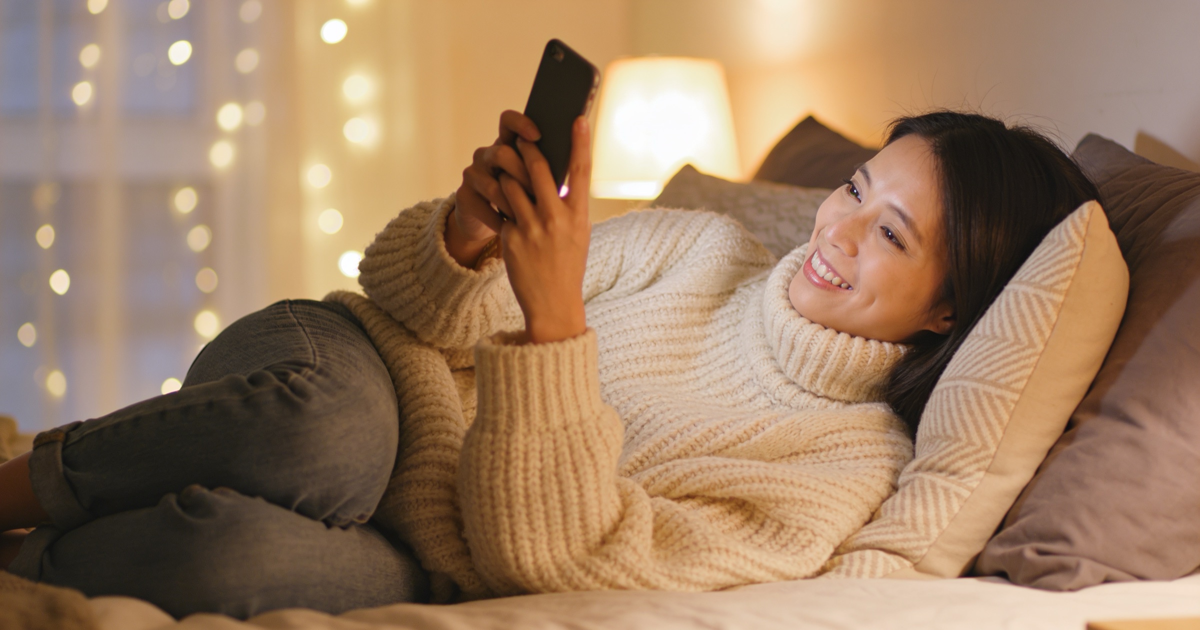 Woman in an oversized knit sweater lying against pillows on a bed, laughing at her phone under warm string lights